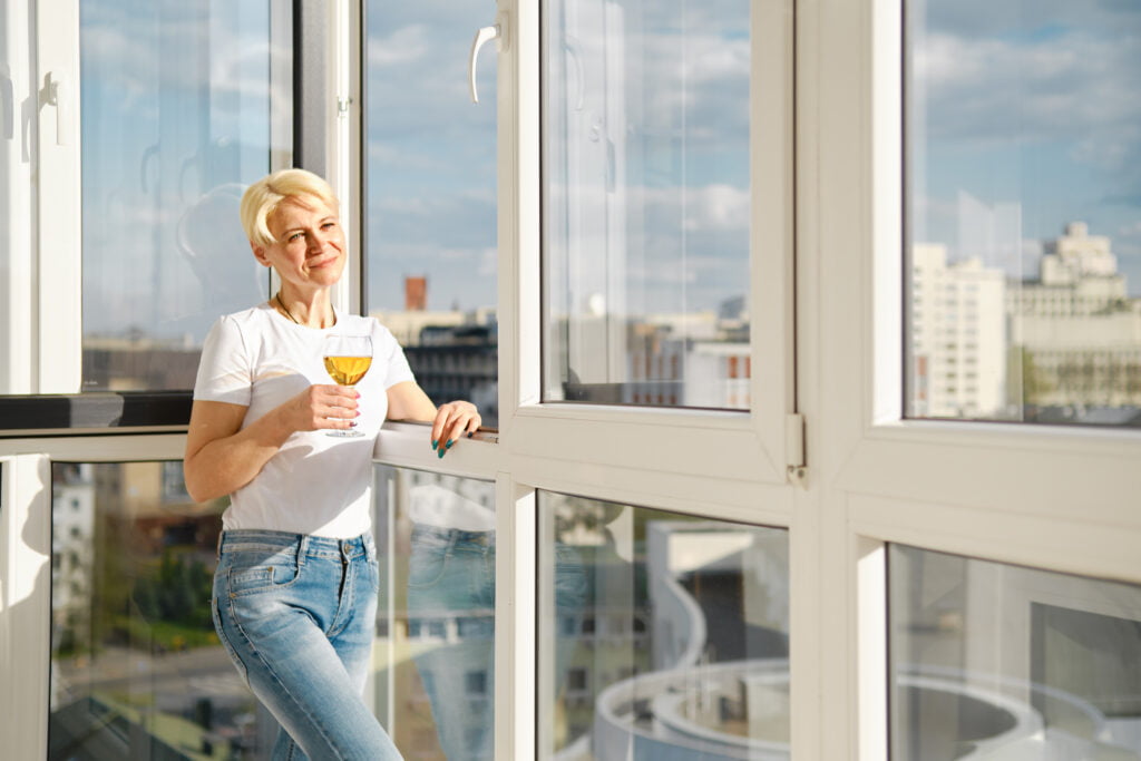 Positive middle-aged woman on balcony of high-rise apartment with glass of juice under bright sun
